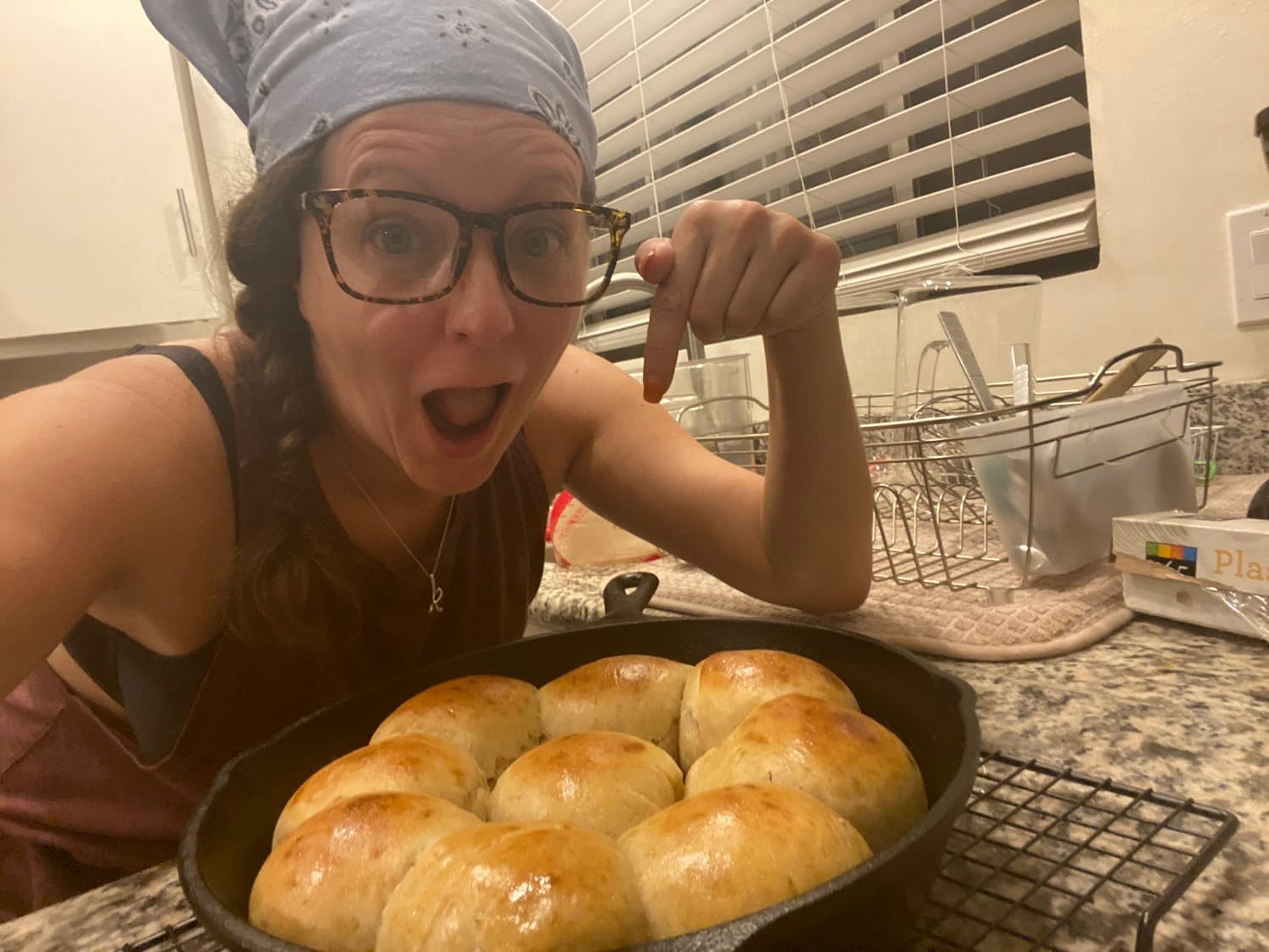 Katie in a bandana, pointing at a cast-iron pan of fresh-baked rolls